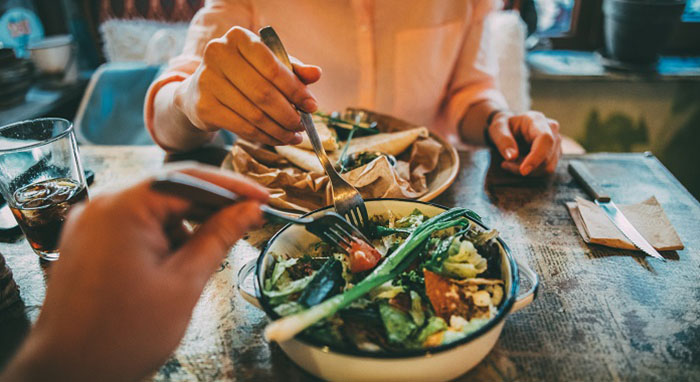 two people eating dinner at a restaurant. All you see is there hands and healthy meals. She's eating pasta and he has a big bowl of vegetables.