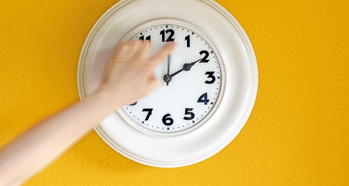 Person reaching out and turning back the time on a clock. It's white against a yellow wall.