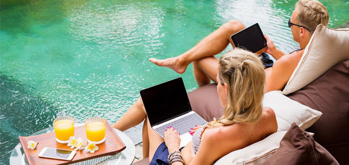 A man and a woman sitting by the pool. Both are on their laptops, with two tropical fruit juice drinks on hand.