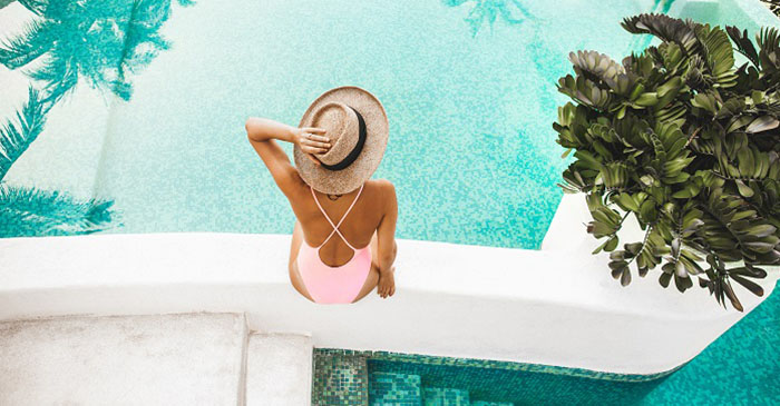 Woman sitting on the wall beside the pool. She's wearing a big straw hat, pink bathers and has her hand stretched up to hold her hat in place.