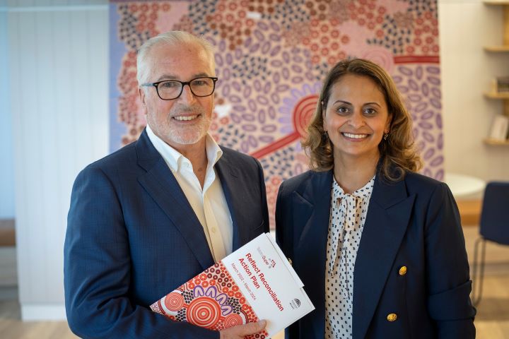 Chris Davies and Krithika Hansen standing in front of an Aboriginal artwork by First Nations Artist Kamara Jones. Chris is holding the TelstraSuper Reconciliation Action Plan. Both are smiling. The artwork features pinks, purples, browns and reds and tells a story of reconciliation.